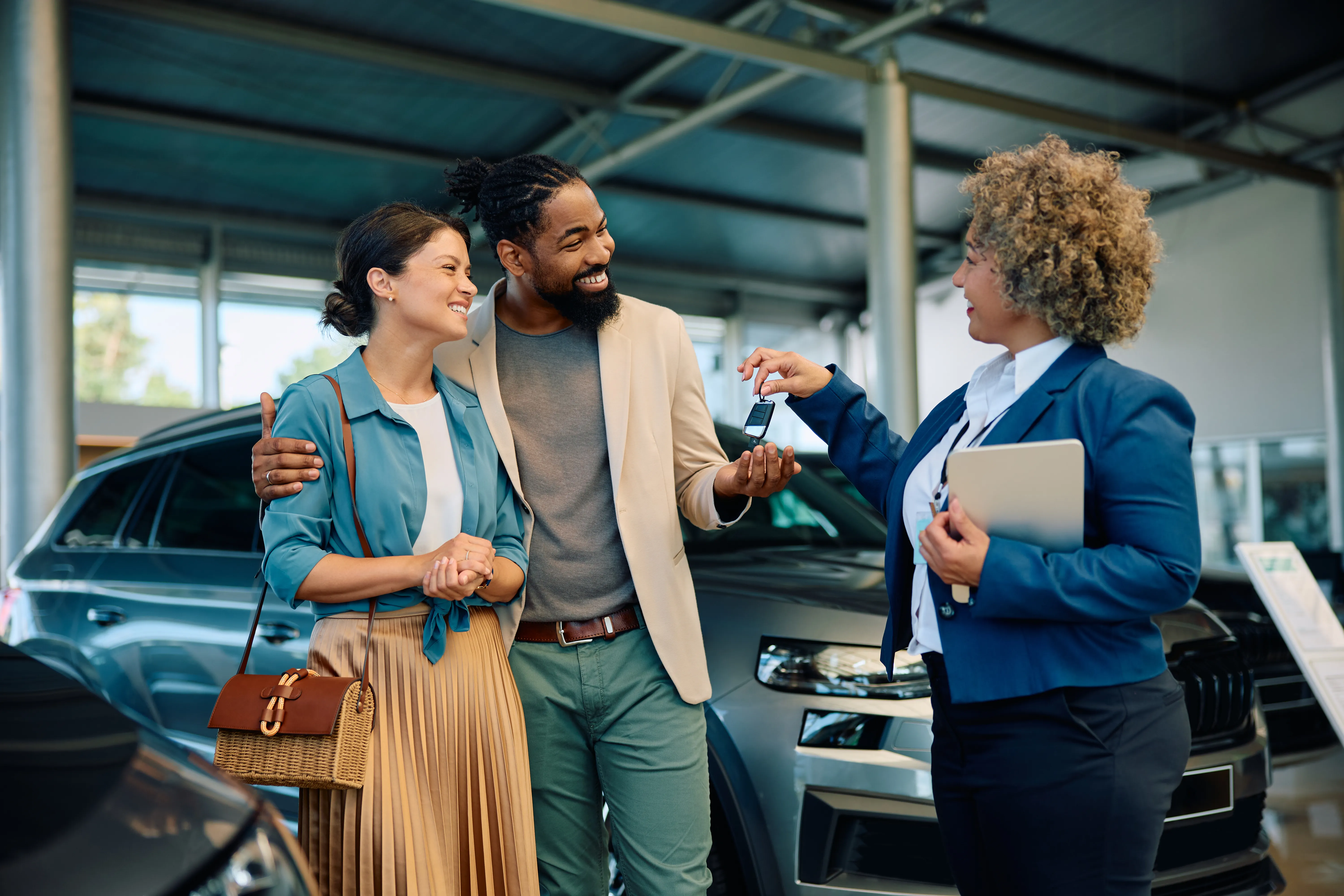 Happy multiracial couple receiving keys of their new car from saleswoman in showroom.